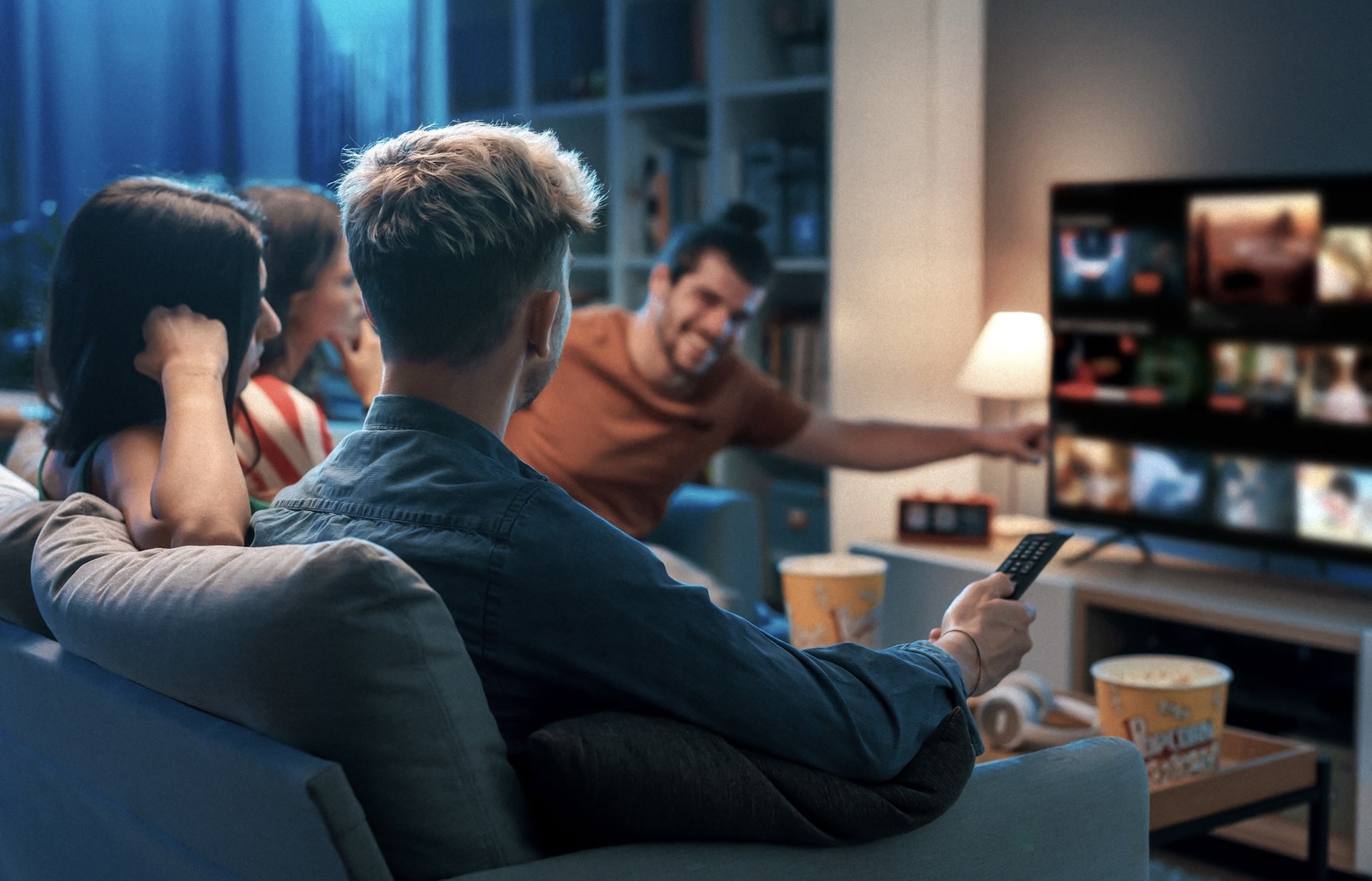 Four people sit on a couch watching a TV screen, with one person holding a remote. Popcorn buckets are on the table in front of them. One person points at the TV screen.